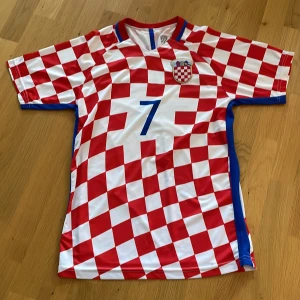 Kroatien fotbollströja  - Säljer en röd och vit fotbollströja med rutmönster och nummer 7 på framsidan. Tröjan har korta ärmar och ett emblem på bröstet. Perfekt för fotbollsfans!
