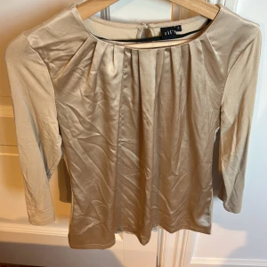 Beige långärmad topp från Rils - Elegant beige topp från Rils med långa ärmar och rund halsringning. Toppen har veckade detaljer framtill och en knappdetalj i nacken. Perfekt för en stilren look.