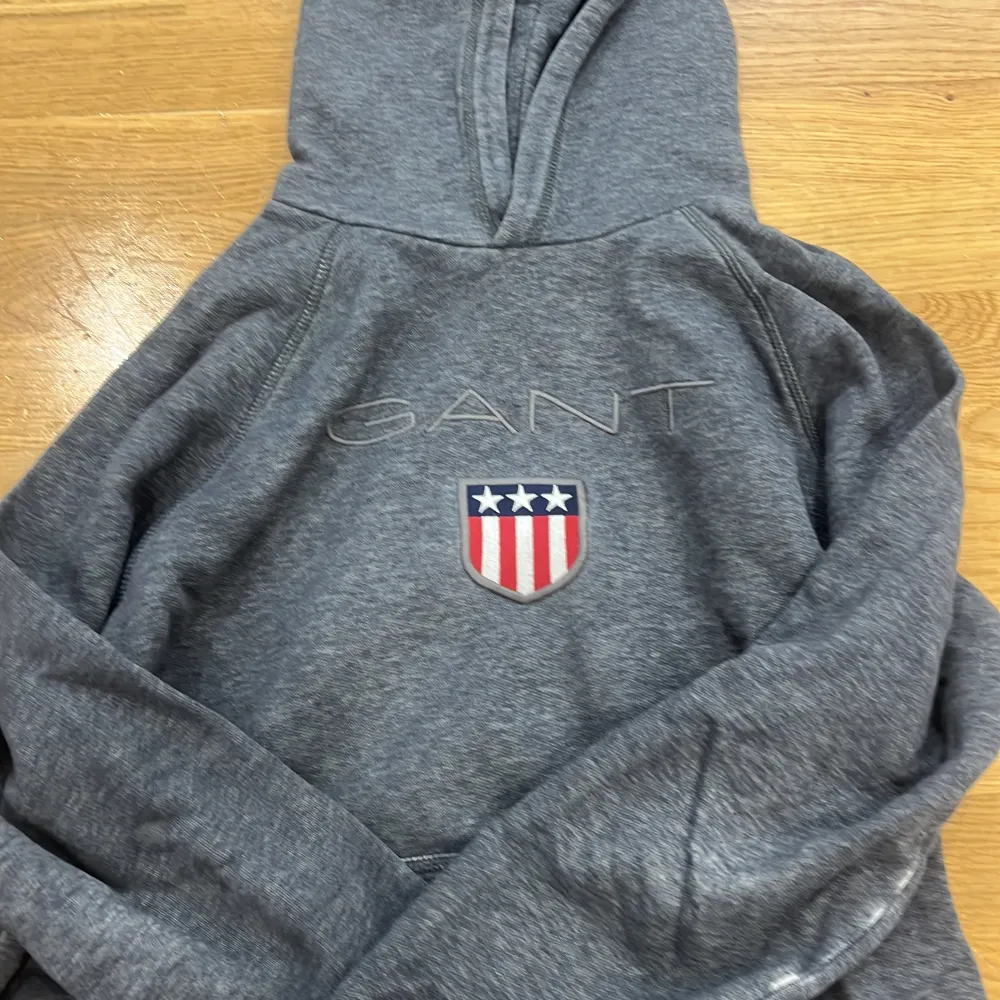 Säljer en stilren grå hoodie från GANT med broderad logga och emblem på bröstet. Tröjan har långa ärmar och en klassisk känguruficka framtill. Perfekt för en avslappnad look.. Neuletakit & Villapaidat.
