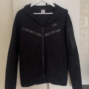 Svart Nike Tech Fleece - Säljer en svart Nike Tech Fleece. Stilren design med en diskret logga på bröstet och en praktisk ficka med dragkedja på armen. Köpt på JD. Nypris: 1349 kr.