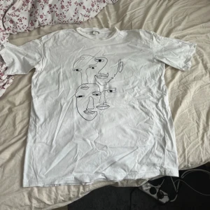 Vit t-shirt med konstnärligt tryck från Monki - Säljer en vit t-shirt från Monki med ett coolt konstnärligt ansiktstryck i svart. Perfekt för dig som gillar unik design och vill sticka ut. T-shirten är kortärmad och gjord i mjuk bomull. Passar perfekt till jeans eller kjol för en avslappnad look.