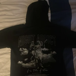 Svart hoodie från Night Addict - Säljer en svart hoodie från Night Addict med tryck på baksidan. Hoodien har en cool design med texten 'Night Addict' och ett konstnärligt motiv. Perfekt för en avslappnad stil. Den är xs men passar lika bra på s 
