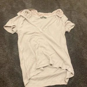 Beige kortärmad topp från Zara - Säljer en stilren beige topp från Zara med korta ärmar och v-ringning. Toppen har knappar framtill och är tillverkad i ett mjukt material. Perfekt för en avslappnad look.