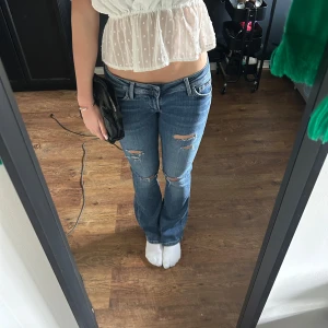 Blå slitna jeans - Säljer ett par blå jeans med slitningar på knäna och låren. De har en klassisk femficksdesign och en låg midja. Perfekta för en avslappnad stil.