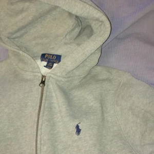 Grå hoodie från Ralph Lauren - Säljer en klassisk grå hoodie från Ralph Lauren med dragkedja och det ikoniska blå logotypbroderiet på bröstet. Perfekt för en avslappnad stil. 🐎