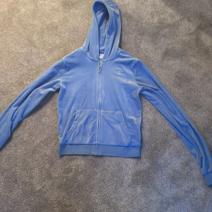 Blå hoodie från Juicy Couture - Säljer en snygg blå hoodie från Juicy Couture. Den är i mjukt material med dragkedja och fickor framtill. Perfekt för en avslappnad stil. Den har en broderad logga och en bekväm huva.