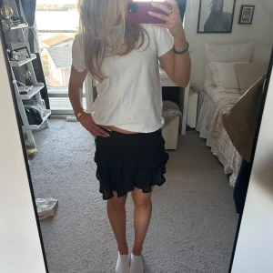 Svart kortkjol med volang - Säljer en svart kortkjol med volangdetaljer. Kjolen har en lekfull design och är perfekt för en trendig look. Passar bra till både vardag och festliga tillfällen.