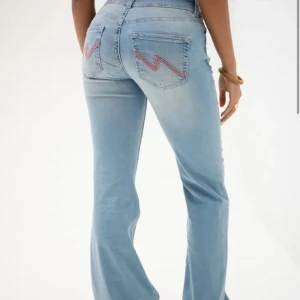 Ljusblå bootcut jeans med broderad detalj - Snygga ljusblå bootcut jeans med låg midja och coola broderade detaljer i rosa på bakfickorna. Jeansen har en klassisk femficksdesign och stängs med två knappar framtill. Perfekta för en avslappnad stil med en twist.