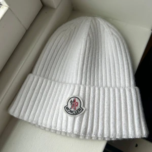 Vit ribbad mössa från Moncler - Snygg vit ribbad mössa från Moncler med broderad logotyp framtill. Perfekt för att hålla värmen under kyliga dagar. Denna mössa är både stilren och funktionell, ett måste i vintergarderoben. Kontakta vid frågor eller bilder