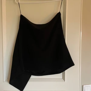 Svart topp från BikBok - Snygg svart asymmetrisk topp från BikBok med tunna axelband. Perfekt för en stilren look. Toppen har en unik design med en längre sida som ger en cool twist. Den har använts en gång och är i storlek M