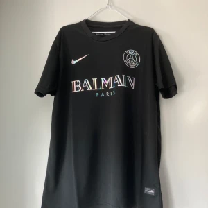 Svart PSG x Balmain fotbollströja - Säljer en helt ny svart PSG x Balmain fotbollströja från Nike. Tröjan har korta ärmar och ett stilrent tryck med Balmain Paris och PSG-loggan i regnbågsskimrande färger. 