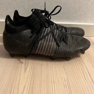 Svarta fotbollsskor i Elite - Säljer ett par svarta fotbollsskor med snörning och ribbad struktur. Skorna har en strumpliknande passform och är designade för att ge bra grepp på planen. Dem är lite trasiga i strumpan. 