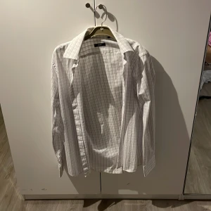 Vit rutig skjorta från Hugo Boss - Säljer en stilren vit skjorta från Hugo Boss med ett diskret rutmönster i blått och rött. Skjortan har långa ärmar och klassisk krage, perfekt för en elegant look. Passar bra till både formella och avslappnade tillfällen.