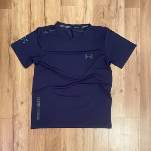 Mörkblå t-shirt från Under Armour - Snygg mörkblå t-shirt från Under Armour med korta ärmar och logga på bröstet. Perfekt för träning med sitt lätta och bekväma material. Diskret design med märkesnamn längs sidan.