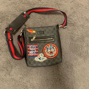 Axelväska från Gucci med patchar - Snygg axelväska från Gucci i grått med coola patchar, inklusive en tiger och en orm. Väskan har en justerbar axelrem i rött och blått samt en praktisk dragkedja framtill. Perfekt för att ge din outfit en unik touch.