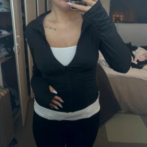 Svart träningsjacka med dragkedja - Säljer en svart träningsjacka med dragkedja framtill. Jackan har en tight passform och långa ärmar med tumhål. Perfekt för träning eller en aktiv vardag.