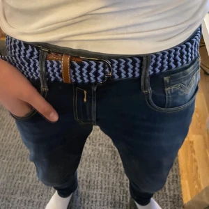 Blått flätat bälte - Snyggt blått flätat bälte med bruna skinn-detaljer och silverfärgat spänne. Perfekt för att ge en stilren touch till din outfit. Bältet har en elastisk design som gör det bekvämt att bära.