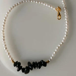 Halsband med pärlor och svarta stenar - Elegant halsband med vita pärlor och svarta stenar i mitten. Det har en guldfärgad låsdetalj som ger en lyxig touch. Perfekt för att lyfta vilken outfit som helst. Handgjort för kvalité!