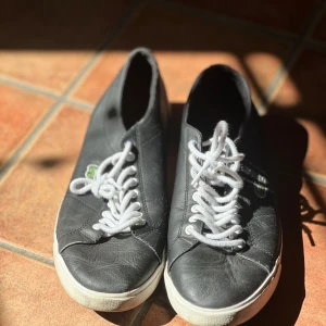 Svarta sneakers från Lacoste - Svarta sneakers från Lacoste med klassisk låg profil och rund tå. Skorna har vit platt sula och vita snören, samt den ikoniska gröna krokodilloggan på sidan. Tillverkade i skinn för en clean och stilren look som funkar till det mesta.