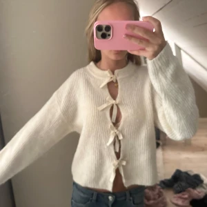 Vit stickad kofta med knyt - Superfin vit stickad kofta med croppad passform och långa ärmar. Koftan har flera söta knytband framtill som ger en unik look. Ribbad struktur och mjukt material, perfekt att styla med jeans eller kjol. Fräsch och trendig design för dig som gillar detaljer.