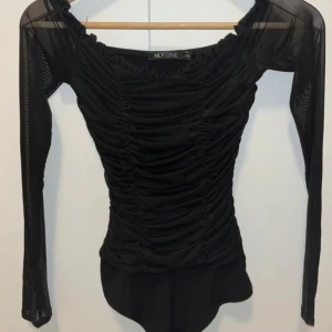 Svart mesh bodysuit från NLY ONE XS - Snygg svart bodysuit från NLY ONE i storlek XS. Toppen har meshmaterial, långa ärmar och är tight i passformen. Rynkad design framtill och runt halsen ger en cool look. Perfekt för att lyfta din outfit med lite edge.