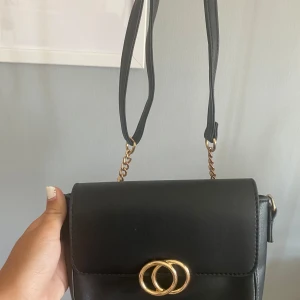 Svart axelväska med gulddetaljer - Snygg svart axelväska från H&M med guldfärgad kedja och dekorativa ringar på locket. Väskan har ett stilrent, fyrkantigt format och är tillverkad i skinnimitation. Insidan har fack och dragkedja för smart förvaring. Perfekt för alla.