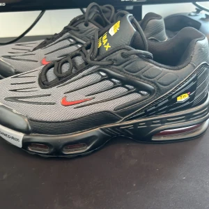 Nike Air Max Plus svarta/grå sneakers - Nike Air Max Plus sneakers i svart och grå mesh med röda swoosh-detaljer och gula TN Air Max-loggor på plösen. Snyggt mönstrad ovandel med svarta plastdetaljer och robust, synlig luftdämpad sula. Skorna har snörning och rund tå, perfekt för dig som gillar streetwear.