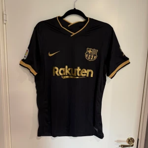 Svart Barcelona fotbollströja  - Snygg FC Barcelona matchtröja från Nike i svart med guldiga detaljer och loggor. Kortärmad med V-ringad krage och Dri-Fit material som andas. Rak passform, guldiga tryck från Rakuten, UNICEF och Beko samt La Liga-märke på ärmen. Perfekt för dig som älskar Barca!