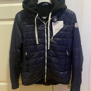 Moncler Vintage Jacka - OBS: Hoodie under jackan, storlek 3 sitter som M. Snygg mörkblå pufferjacka från Moncler med quiltad design och vita snören. Jackan har flera dragkedjor, huva och Moncler-logga på ärmen. Perfekt för dig som vill ha en stilren och sportig look under kyliga dagar.