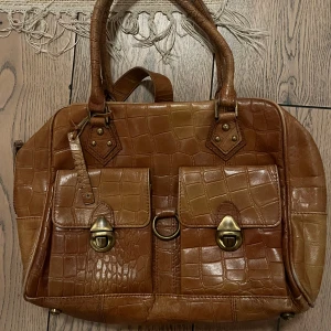 Brun vintage handväska - Snygg brun handväska med metall detaljer. Väskan har två framfickor med metallspännen, dubbla handtag och en avtagbar axelrem. Insidan är rymlig med flera fack och dragkedja. Perfekt för dig som gillar klassisk och cool stil.