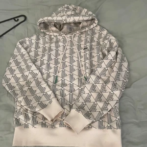 Lacoste hoodie - Säljer en vit hoodie från Lacoste med ett fett grafiskt mönster i svart över hela tröjan. Klassisk huva med snörning och ribbade muddar. Ikoniska Lacoste-loggan på bröstet. Perfekt för dig som gillar streetwear och vill sticka ut.