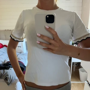 Vit topp med spetsdetalj från Zara - Supersöt vit topp från Zara med korta ärmar och unika virkade spetsdetaljer i svart, vitt och guld vid ärmsluten. T-shirten har en klassisk rund halsringning och är gjord i mjukt bomullsmaterial. Perfekt för dig som gillar stilrena plagg med en twist!