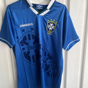 Blå Brasilien Romario 11 Umbro tröja - Säljer en blå Brasilien fotbollströja från Umbro med Romario och nummer 11 på ryggen. Tröjan har korta ärmar, vit och grön detalj vid kragen, samt stora CBF-loggor och stjärnor på framsidan. Materialet är lätt och ventilerande polyester, perfekt för match eller träning.