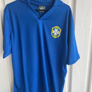 Blå Brasilien Pelé 10 fotbollströja - Snygg blå fotbollströja med Brasilien CBD-emblem på bröstet och klassisk krage. På ryggen står det 'Pelé' och nummer 10 i vitt. Tillverkad i mjukt, lätt syntetmaterial som andas. Perfekt för dig som vill hylla en legend på planen eller på läktaren.