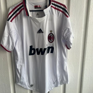 AC Milan Ronaldinho 80 Adidas tröja - Säljer en vit AC Milan fotbollströja från Adidas med Ronaldinho och nummer 80 på ryggen. Tröjan har korta ärmar, röda och svarta detaljer på axlarna och kragen, samt Champions League-patch på ärmen. Materialet är lätt och andas, perfekt för match eller träning.