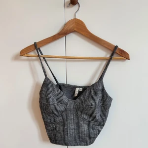 Grå korsett-topp - En grå korsett-topp från Nelly i grått med rutigt mönster och smala axelband. Toppen har markerade kupor och en figurnära passform.