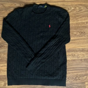 Svart stickad tröja Polo Ralph Lauren - Svart kabelstickad tröja från Polo Ralph Lauren med klassisk röd broderad logga på bröstet. Rund halsringning och ribbade muddar vid ärmslut och nederkant. Perfekt för dig som gillar stilren och tidlös design.