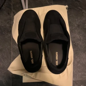 Svarta sneakers från Axel Arigato - Snygga svarta sneakers från Axel Arigato med minimalistisk design och rund tå. Skorna har elastiska paneler för enkel påtagning och är tillverkade i mocka och skinn. Perfekta för dig som gillar stilrena och moderna skor.