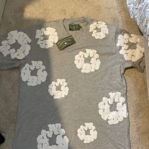 Grå blommig t-shirt från Denim Tears - Snygg grå t-shirt från Denim Tears med vita blommiga tryck över hela plagget. T-shirten har rund hals och korta ärmar, tillverkad i mjuk bomull som är skön mot huden. Perfekt för dig som gillar streetwear med unik design. mått B 48 Cm/ L 71 Cm