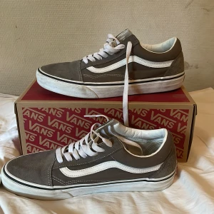 Grå Vans Old Skool sneakers - Säljer ett par grå Vans Old Skool sneakers med vit sula och klassisk vit sidostripes. Skorna har snörning och rund tå, tillverkade i mocka och syntet. Perfekt för dig som gillar streetstyle och vill ha en ikonisk look.