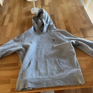 Grå hoodie från Polo Ralph Lauren - Säljer en grå hoodie från Polo Ralph Lauren med klassisk huva och stor magficka. Tröjan har lång ärm och den ikoniska lilla Polo-loggan broderad på bröstet. Perfekt för chill dagar eller när du vill ha en enkel och snygg look.   STORLEK 176