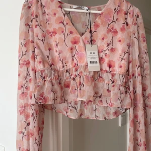 Blommig rosa blus från NA-KD - Supersöt blommig blus från NA-KD i ljusrosa toner med v-ringning och croppad passform. Blusen har långa ärmar med knyt vid mudden och volangdetaljer vid midjan. Kan knytas/spännas åt vid midjan 
