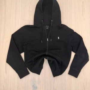 Svart croppad hoodie från Polo Ralph Lauren - Svart croppad hoodie från Polo Ralph Lauren med vit broderad logga på bröstet. Tröjan har huva med snörning, dragkedja framtill och långa ärmar. Perfekt för dig som gillar en chill och stilren look.