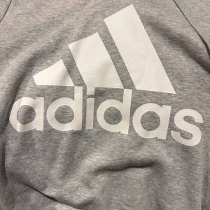 Grå Adidas hoodie med stor logga - Snygg grå hoodie från Adidas i storlek XS med stor vit logga på bröstet. Tröjan har huva med snörning, ribbade muddar och känguruficka framtill. Perfekt för chill eller träning, mjukt material i bomull och polyester.