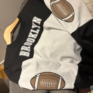 Brooklyn sweatshirt med fotbollstryck - Cool svart och vit sweatshirt med BROOKLYN-tryck över bröstet och stora amerikanska fotbollar på ärmarna. Tröjan har breda blockränder och rund hals. Materialet känns som mjuk bomull eller polyester, perfekt för chill dagar eller sportig stil.