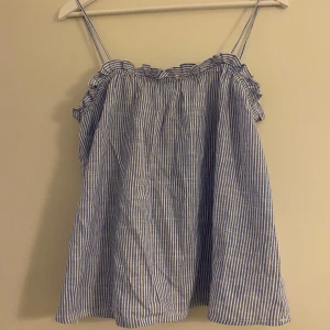 Randig blå linnetopp från H&M XS - Säljer en söt och luftig linnetopp från H&M i storlek XS. Toppen har tunna axelband, volangdetaljer vid halsringningen och ett blåvitt randigt mönster. Perfekt för varma dagar och enkel att matcha med jeans eller shorts.