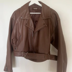 Brun skinnjacka från Bianca x Nelly - Säljer en snygg brun skinnjacka från Bianca x Nelly med coola detaljer som bred krage, dragkedja och bälte i midjan. Jackan har lång ärm och en loose passform som ger en chill vibe. Perfekt för dig som vill ha en trendig look med edge.