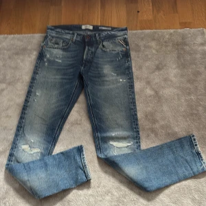 Replay Grover blå jeans med slitningar - Säljer ett par Replay willbi jeans i klassisk blå tvätt med snygga slitningar och detaljer på bakfickorna. Jeansen har rak passform och normal midja, tillverkade i robust denim. Perfekt för dig som gillar en avslappnad och cool stil.