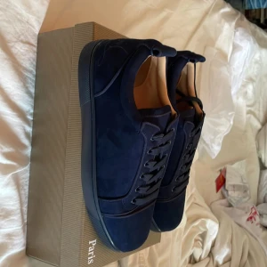 Mörkblå sneakers från Christian Louboutin - Exklusiva mörkblå sneakers från Christian Louboutin i mocka med klassisk röd sula. Snygg och stilren design med rund tå, platt sula och snörning. Perfekta för dig som vill sticka ut med lyxiga detaljer och hög modefaktor.