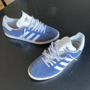 Adidas Gazelle blå mocka sneakers - Säljer ett par Adidas Gazelle sneakers i blå mocka med vita detaljer och klassiska tre ränder på sidan. Skorna har vit platt sula, rund tå och snörning. Tungan och hälen är i vitt syntetmaterial med Adidas-logga. Perfekt för dig som gillar retrostil och streetwear.
